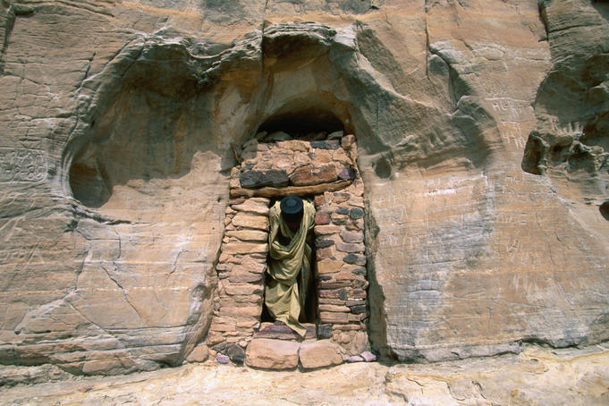 A priest exits through the stone doorway in a rock cliff face; the main entrance to the well-concealed church of Abuna Yemata, near Hawsien