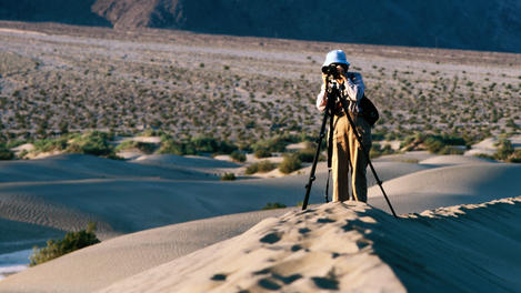 Woman standing on dune taking photo with camera on tripod.