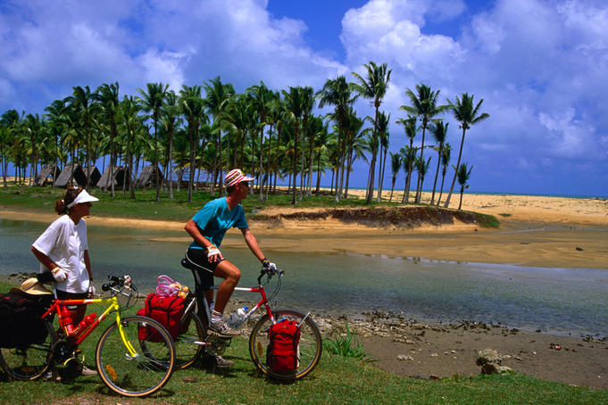 Cyclists rest at a beach on the East Coast of Malaysia with a backdrop of palm trees and thatched huts.