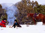 Cowboy with cowgirl sitting near camp fire in snow, their horse standing nearby.