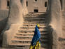 A local woman walking past the entrance to the mosque built in the traditional Sahel style, using mud bricks and wooden beams that extend from the walls so that ladders can be supported when it comes time to repair the mud rendering after the rainy season - Djenne, Mopti
