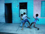 Three boys playing with a football on the pavement outside a row of painted buildings