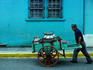 A milkman, pushing a painted wooden cart loaded with milk cans, past a painted wall