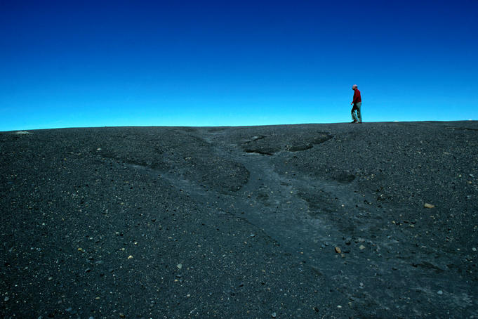 Man walking on rim of Irazu volcano crater, against blue sky.