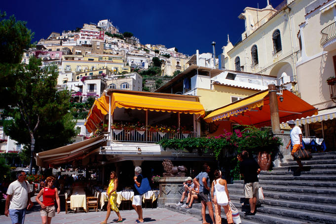 Tourists near waterfront restaurants with hillside houses in background.