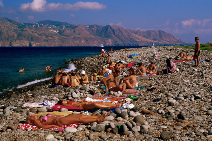 Sunbathers on stone beach with Lipari in background.