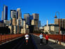 Women jogging across Stone Arch bridge with city skyline beyond.
