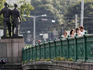 People crossing green bridge decorated with socialist statues connecting old and new towns.