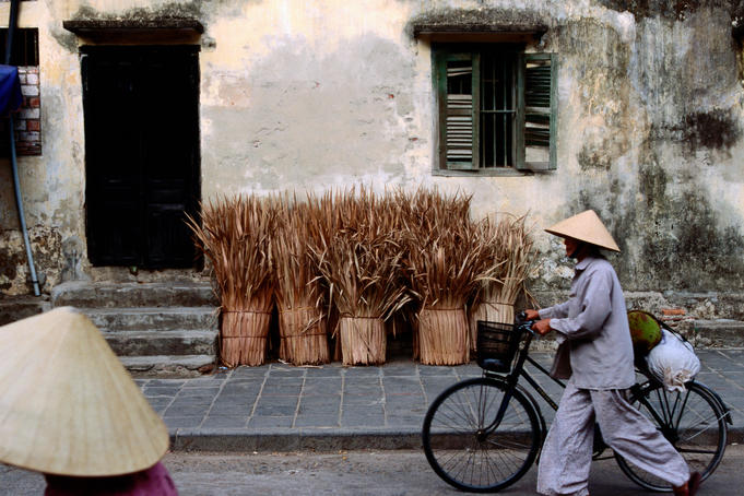 Person wearing conical hat walking bike down street past weathered building and bunches of straw.