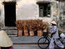Person wearing conical hat walking bike down street past weathered building and bunches of straw.