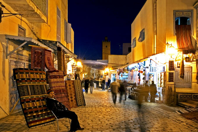 People strolling along cobblestone street past carpet seller at twilight.