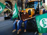 Farmers protesting on tractor bucket in street parade.