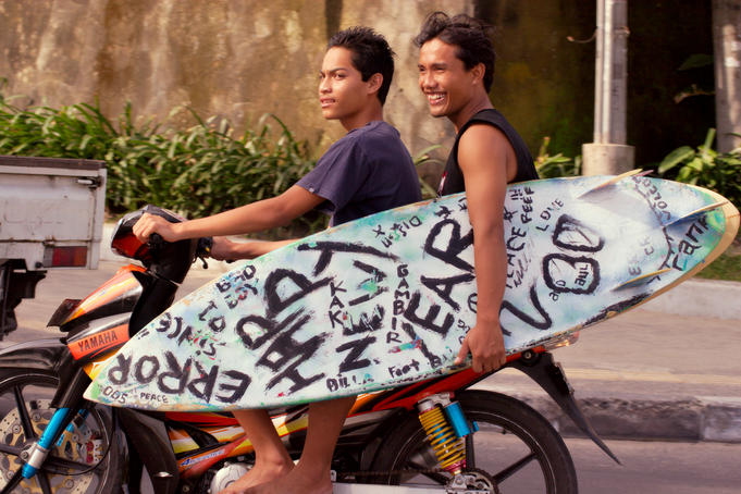 Two locals on motor bike with surfboard.