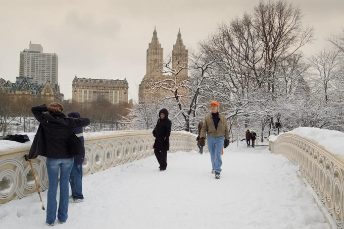 People crossing snow-covered Bow Bridge, linking the Ramble with Cherry Hill, with San Remo apartments in background.