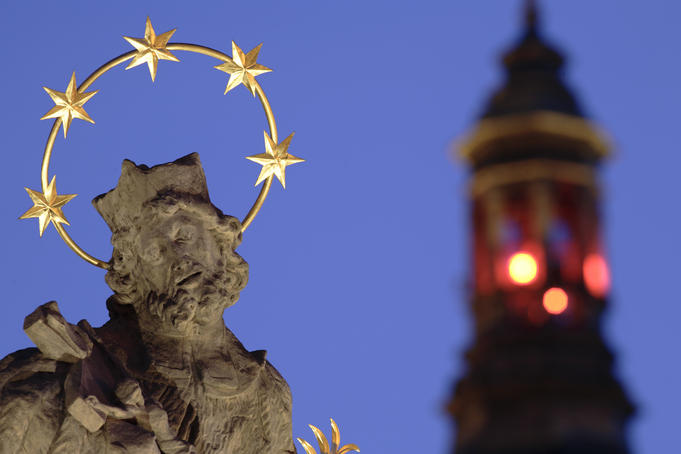 Floodlit statue of saint on plague column against tower with red lights in Velke Namesti Square.