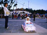 Mother smiling at girl in toy beetle car at dusk, with families behind.