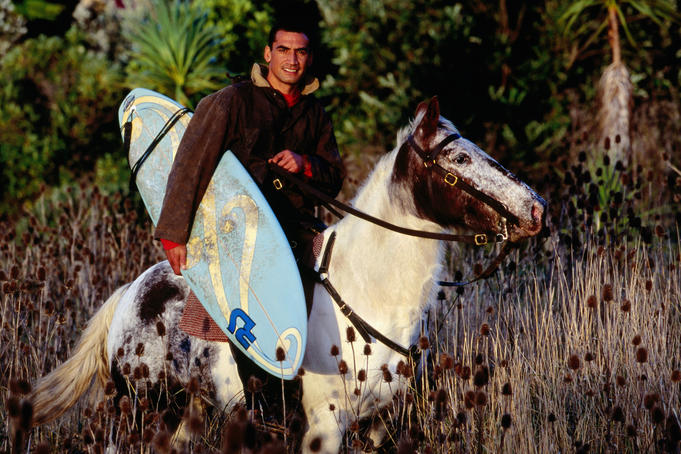 Local Maori surfer carrying his surfboard while on horseback.