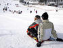 Two people on sled at top of run with people below.