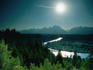 The sun, hanging low in the sky, reflects off the silver ribbon of the Snake River, leading the eye to the Teton Range; viewed from the Snake River Overlook - Grand Teton National Park, Wyoming