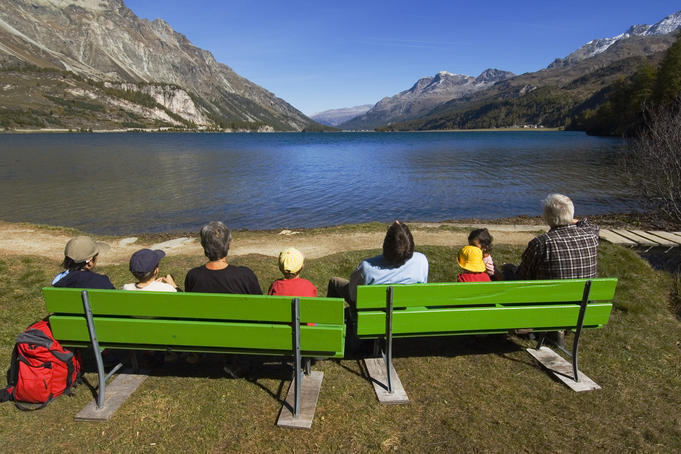 People sitting on green benches at alpine Silser See (lake).