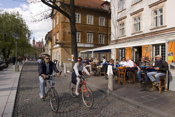 Cyclists passing outdoor cafes along river.