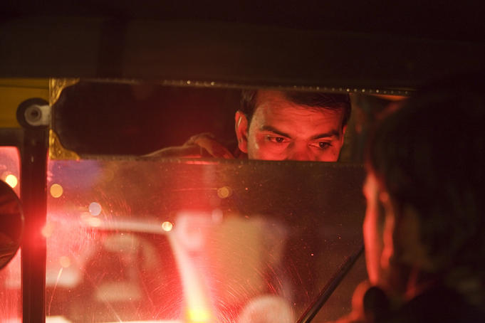 Brake lights reflect off an Auto Rickshaw drivers face.