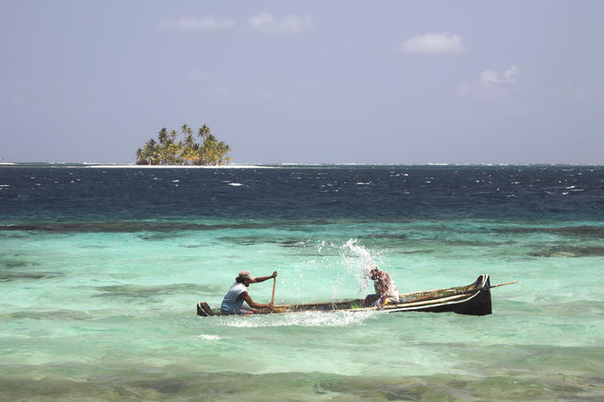 Man paddling while another man bales water out of wooden boat in open water.