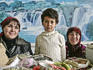 Young girl and two women at the bazaar selling spices with a picture of waterfalls behind them.