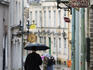 People with umbrellas strolling down narrow brick-laid street in old town Tallinn on rainy afternoon.