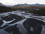 Wide river bed with tangle of streams and snow-covered mountain peaks in background.