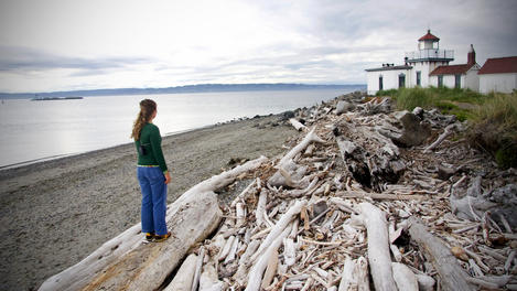 Woman standing on driftwood and looking towards Discovery Park Lighthouse.