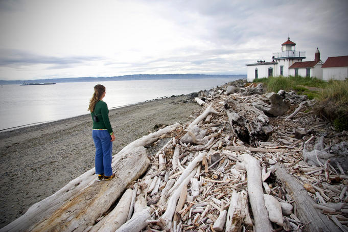 Woman standing on driftwood and looking towards Discovery Park Lighthouse.