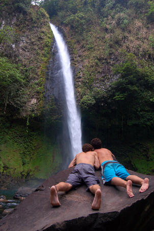 Couple of men laying on rock and viewing waterfall near city of Arenal.