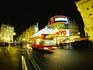 Double decker bus driving past illuminated neon signs of Picadilly Circus at night.