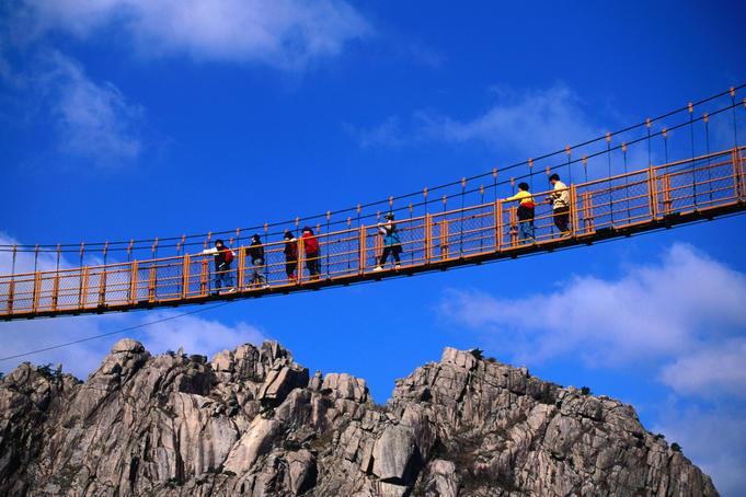 Hikers crossing steel suspension bridge over crevasse.