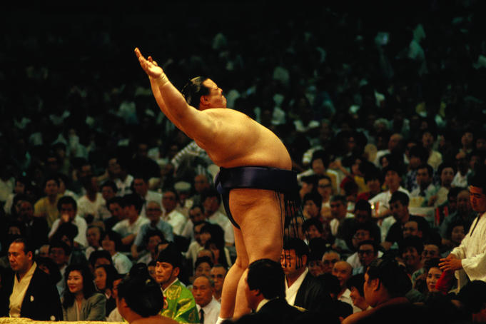 A Sumo wrestler performs his pre-bout stretching to the crowd at a packed stadium.