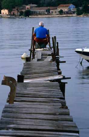A lone angler sitting at the end of a rickety wooden jetty, fishing.