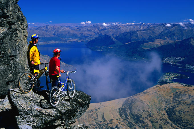 A pair of mountain bikers on top of The Remarkables with a view over Lake Wakatipu and Queenstown.