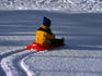 Boy on a red plastic sled going down a small snowy hill.